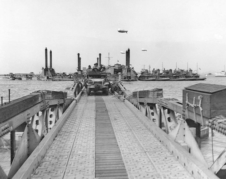 A vehicle drives along the floating roadway of the Mulberry harbour off Omaha Beach; barrage balloons fly over the harbour.