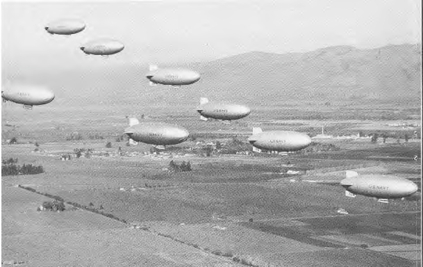 A formation of U.S. Navy blimps in flight over farmland and hills.